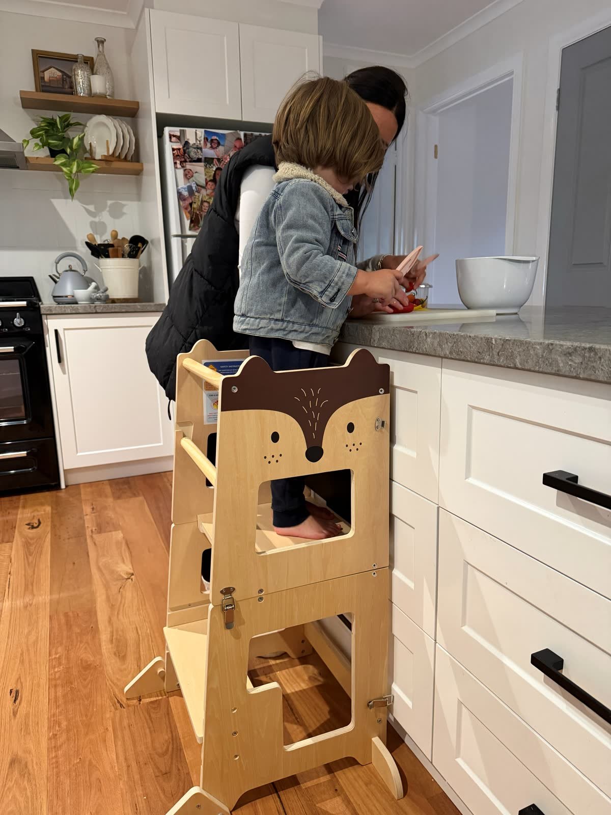 A child standing in a timber learning tower beside an adult at a kitchen bench with fruit and a bowl.
