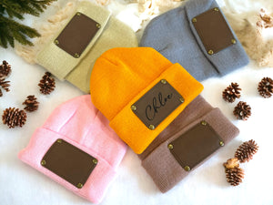 Four beanies in different colors with leather patches on a white background with pine cones.