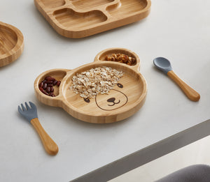 A bamboo bear-shaped plate filled with oatmeal, beans, and nuts, arranged in separate compartments. The plate features a cute bear face design and is accompanied by matching bamboo utensils with gray silicone tips.