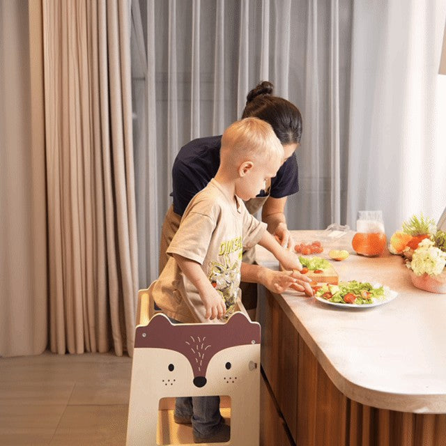 toddler standing at fox learning tower helping at the kitchen counter with an adult