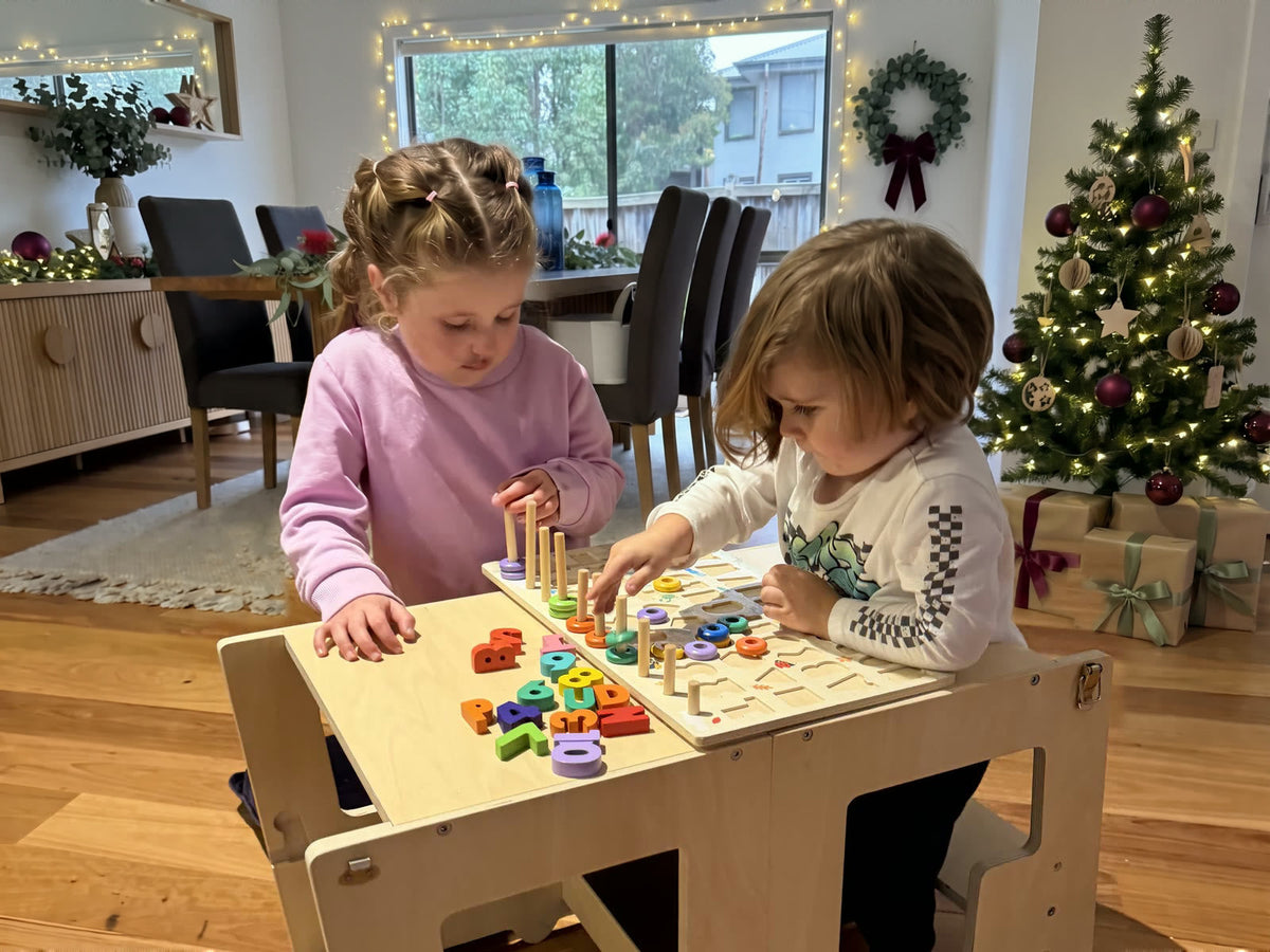 Two young children playing with a wooden number puzzle at a timber Fox learning tower set up as a table and chair, in a room decorated for Christmas with a tree, wrapped presents, a eucalyptus wreath and fairy lights