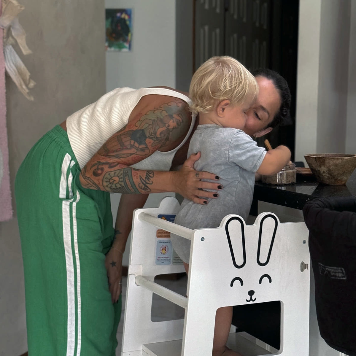 A parent leaning in to hug a toddler who is standing in a white bunny learning tower at a kitchen bench