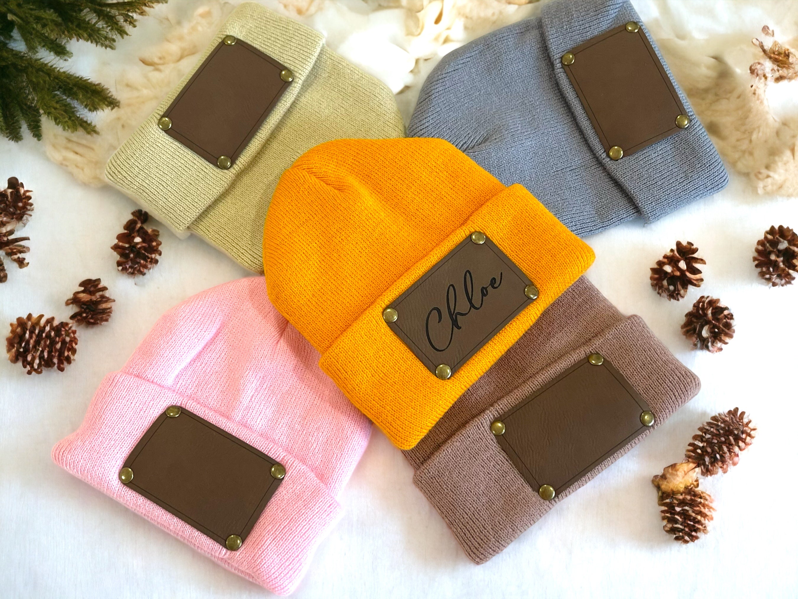 Four beanies in different colors with leather patches on a white background with pine cones.
