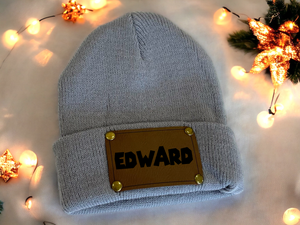 Gray knit beanie with 'EDWARD' patch on a light background with decorative lights.