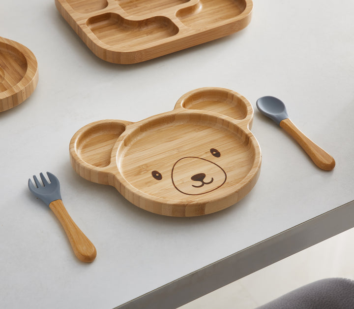 A bamboo bear-shaped plate with empty compartments and a cute bear face design in the center. It is accompanied by matching bamboo utensils with gray silicone tips