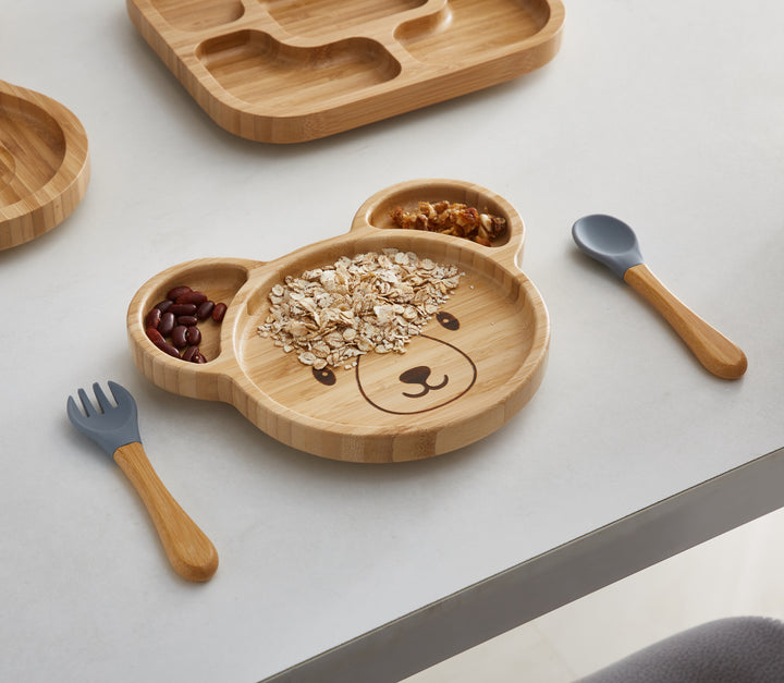 A bamboo bear-shaped plate filled with oatmeal, beans, and nuts, arranged in separate compartments. The plate features a cute bear face design and is accompanied by matching bamboo utensils with gray silicone tips.
