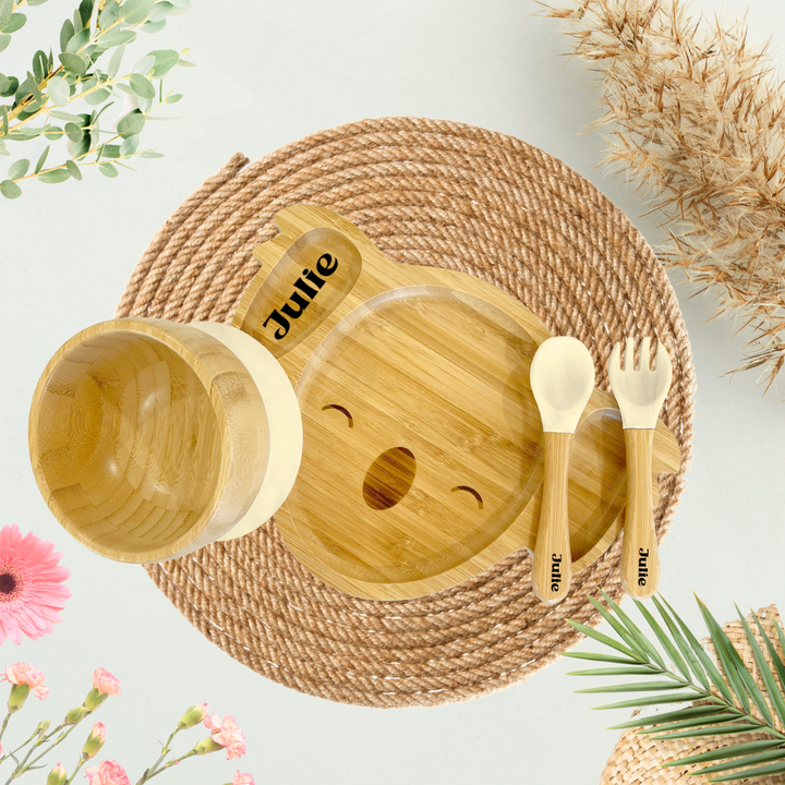 Personalized bamboo koala-shaped plate for toddlers with the name 'Julie' engraved, featuring eco-friendly dining compartments. Includes a matching bamboo bowl and utensils with cream-colored silicone tips, all engraved with 'Julie.' Perfect for sustainable and custom kids' mealtime products is Ideal for promoting environmentally friendly baby and toddler essentials.