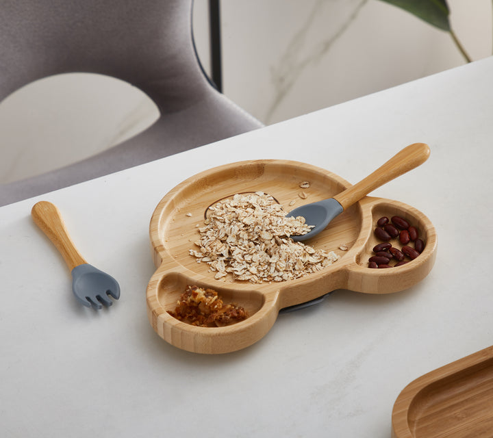 A bamboo bear-shaped plate filled with oatmeal, beans, and nuts, divided into separate compartments. A gray-tipped bamboo spoon rests on the plate, while a matching fork is placed nearby on the light-colored tabletop
