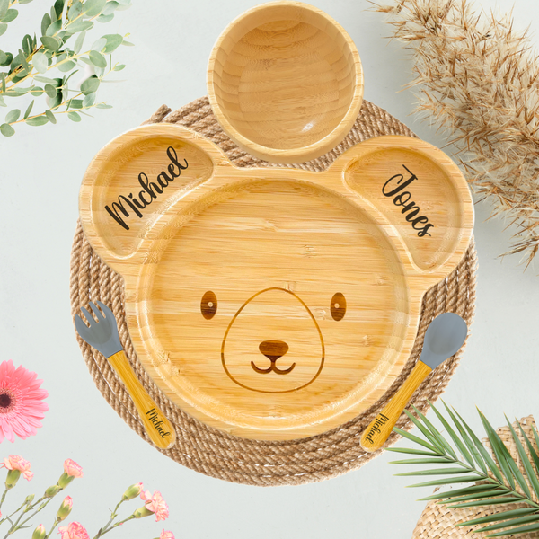 A personalized bamboo plate shaped like a bear, featuring the names "Michael" and "Jones" engraved in the ears and a bear face design in the center. The set includes a small bamboo bowl and matching bamboo utensils with gray silicone tips engraved with the name "Michael." Styled on a round woven placemat, the scene is accented with floral decorations and natural greenery.