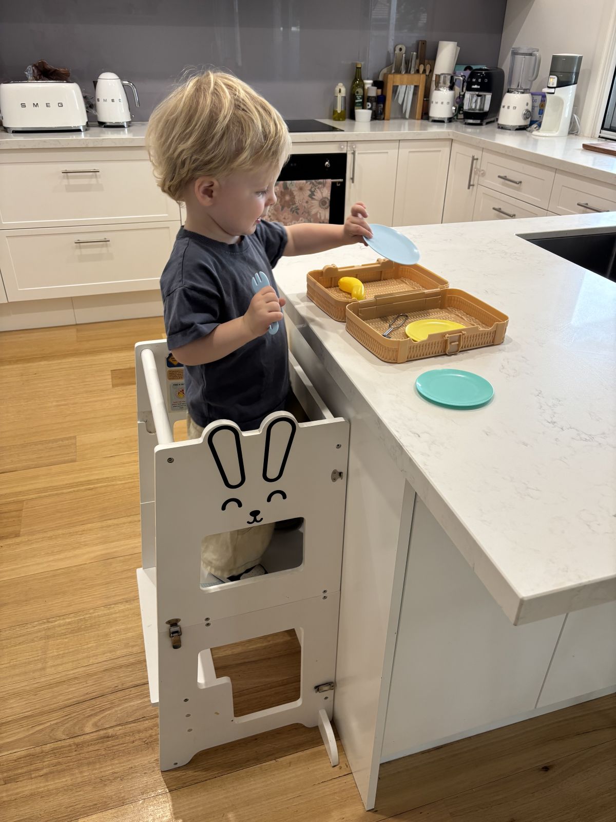 A toddler standing in a white Bunny learning tower at a kitchen island, putting out a blue plate beside two open trays